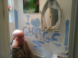 George putting 'crazy finger' to his head in graffitied bathroom
